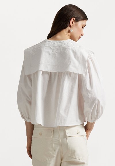 White cotton blouse with a wide collar, puffed sleeves, and subtle floral embroidery on the back. Paired with beige trousers featuring back pockets.