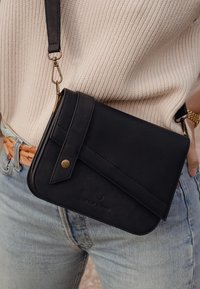 Schwarze Leder-Crossbody-Tasche mit strukturiertem Panel, goldenen Beschlägen und Klappverschluss. Kombiniert mit hellblauen Jeans und beigem Strickoberteil.