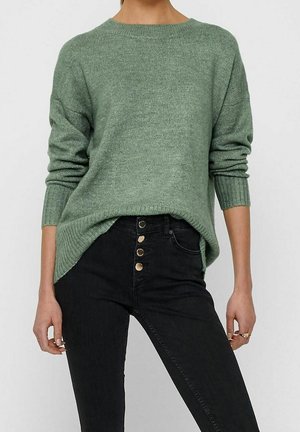Grøn strikket sweater med en afslappet pasform, ribbet halsudskæring og lange ærmer, sat sammen med højtaljede sorte jeans med guldknapper.