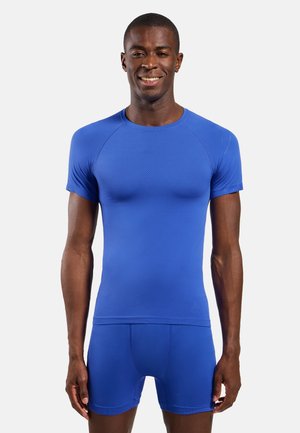 Homme souriant, portant une chemise ajustée bleu vif à manches courtes et un short bleu assorti, debout devant un fond clair uni.