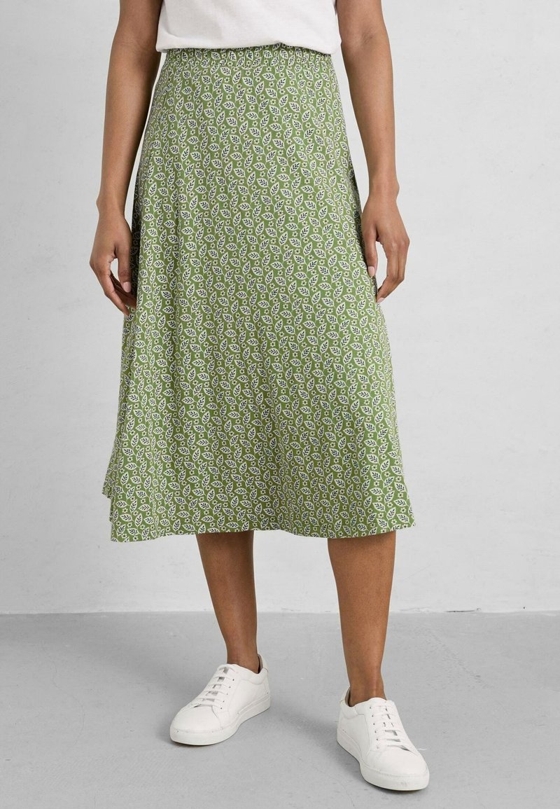Femme portant une jupe midi verte avec un motif floral blanc, des baskets blanches et un haut blanc rentré, debout sur un sol gris.