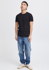 T-shirt en coton noir avec un col rond et des manches courtes, comprenant une poche poitrine. Porté avec un jean en denim bleu clair et des baskets blanches.