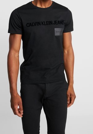 T-shirt en coton noir avec le logo "CALVIN KLEIN JEANS" dans une texture assortie, manches courtes et col rond, associé à un pantalon noir.