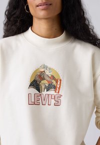 Felpa color crema Levi's con grafica rotonda che raffigura due figure in plaid e denim, accentuate da dettagli rossi e gialli. Texture morbida.