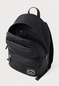 Musta nailonireppu, jossa on useita vetoketjullisia taskuja, säädettävät hihnat ja yläpida. Edessä on brändätty merkki.