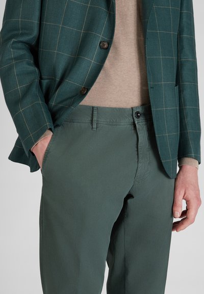 Chinos verdi in cotone abbinati a un blazer verde scuro a fantasia. L'outfit presenta un maglione chiaro sotto e un dettaglio con bottoni sul blazer.