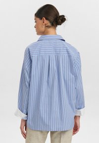Blue and white striped oversized shirt with a pointed collar, button-down cuffs, and a vent at the back. Lightweight fabric texture.