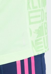 Svijetlozelena sportska majica s mrežastom teksturom, s odvažnim grafičkim motivima i logotipom. Uparen s tamnoplavim šortsem s ružičastim prugama.