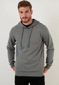 Szürke kapucnis pulóver, amely texturált mintával, bordás részletekkel és laza szabással rendelkezik, állítható zsinórokkal.