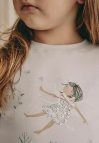 Világos lila pulóver, amelyen egy tündérdesign látható csillogó flitterekkel, pasztell masnival és zöld, kék virágos illusztrációkkal.