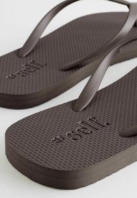 Para ciemnobrązowych teksturowanych klapek z cienkimi paskami i wytłoczonym logo "self." na każdej podeszwie, umieszczonych na jasnej powierzchni.