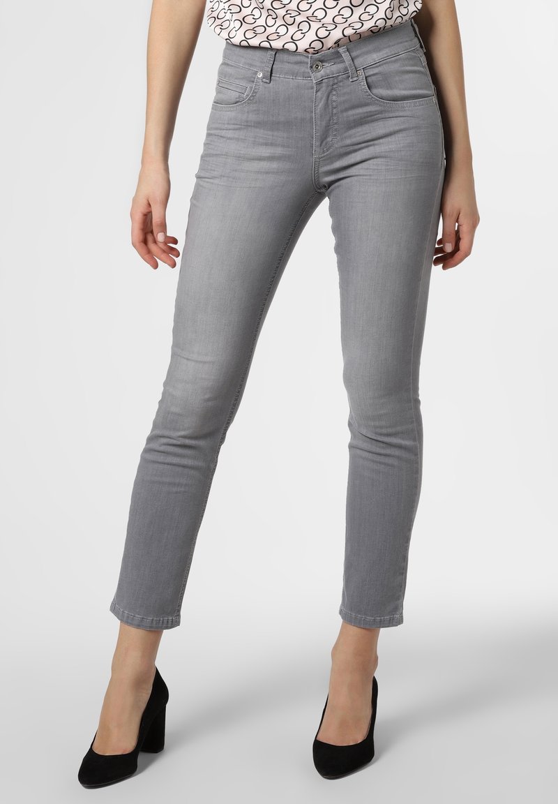 Femme portant un jean gris clair slim et des chaussures à talons noires, debout devant un fond clair uni.