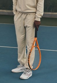 Tennissracket med en orange ram och svart handtag, hålls av en person i beige sweatpants och matchande sweatshirt, som bär vita sneakers.