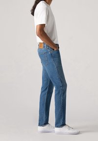 Ljusblå denimjeans med rak passform, med en tan läderpatch bak och klassisk femfickdesign. Bärs med en vit t-shirt.