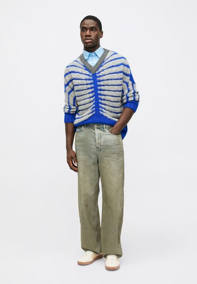 Maglione grigio e blu a righe, con scollatura a V, abbinato a jeans larghi verde oliva e sneaker crema. Camicia blu con colletto sotto.