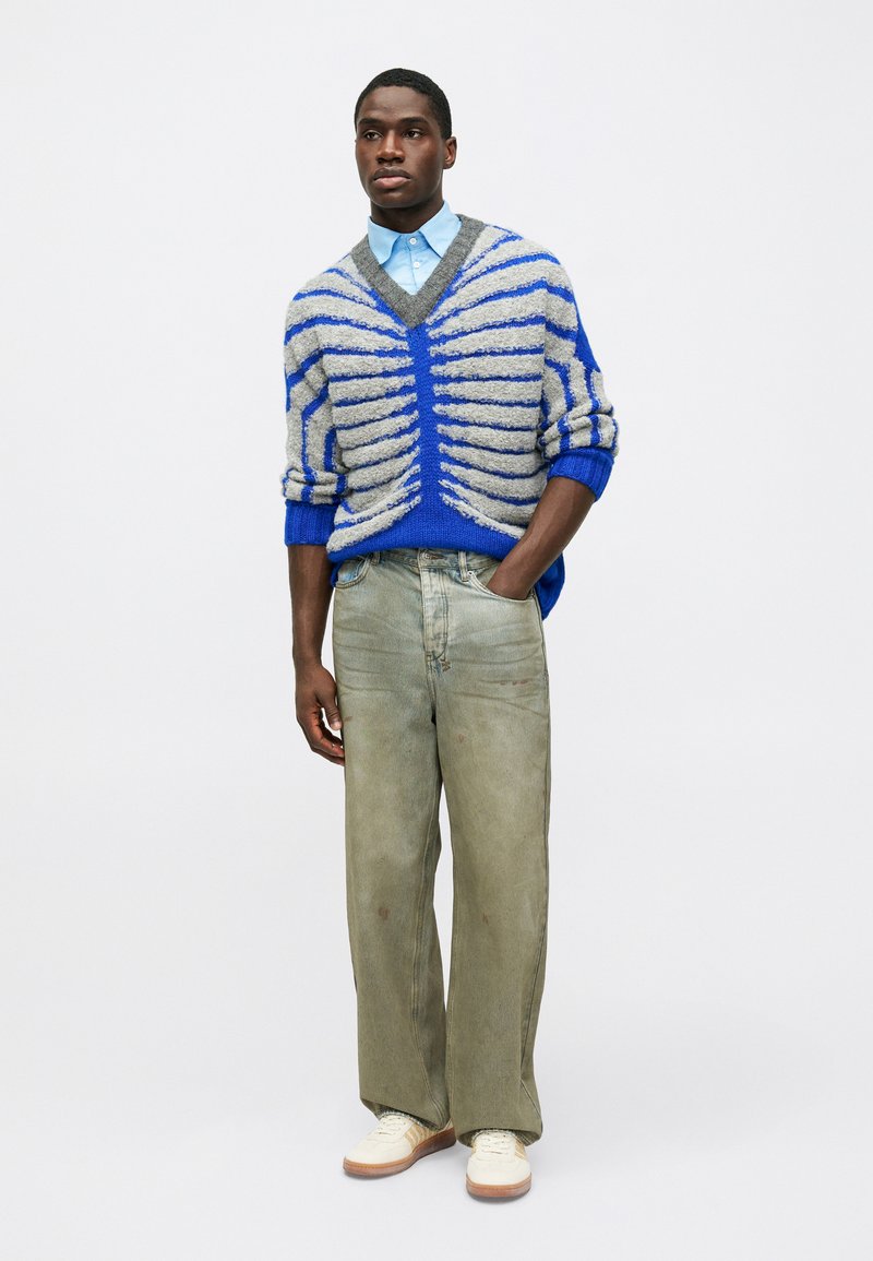 Πλεκτό γκρι και μπλε ριγέ πουλόβερ με V-λαιμό, συνδυασμέν�ο με χαλαρά ελαιόχρωμα τζιν και κρέμα αθλητικά παπούτσια. Μπλε πουκάμισο με γιακά από κάτω.