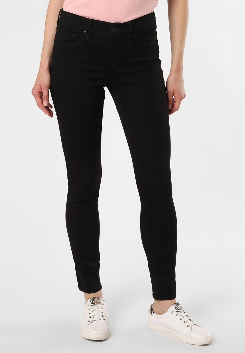 Jeans skinny negros de denim elástico, con cintura media, presillas para cinturón, bolsillos frontales y una textura sutil. Combinados con zapatillas blancas.