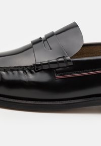 Mocassin en cuir noir avec des détails cousus, une tige surélevée et une accent rouge. La semelle est plate et faite d'un matériau contrastant.