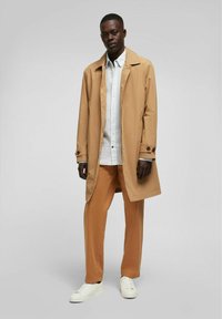 Lichtbeige trenchcoat, enkelvoudig knoop, gedragen over een witte overhemden en oranje broek, gecombineerd met witte sneakers.