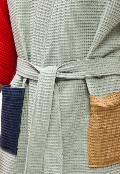 Ανοιχτό γκρι ρόμπα με υφή βάφλας, δεμένη στη μέση, με μια μπλε σκούρα και μια ταμπά τετράγωνη τσέπη, φορεμένη πάνω από κόκκινο ένδυμα.