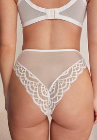 Hvid blondelingeri med en gennemsigtig mesh-ryg, bølget blondekant og en blød tekstur samt et højskåret ben-design.