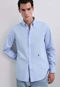 Chemise bleu clair à manches longues en coton, ornée d'un petit emblème de crâne marine sur la poitrine gauche, avec un design de col classique.