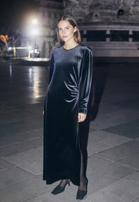 Longue robe droite en velours noir avec manches longues, fronces latérales et fente sur le côté, associée à des escarpins noirs pointus. Cadre extérieur éclairé.