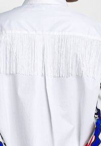 Witte shirt met lange witte franjes aan de achterkant. Het schoudergebied heeft kleurrijke patronen aan de zijkanten. Overal een gladde textuur.