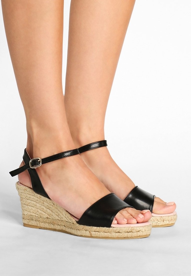 Pieds portant des sandales compensées en cuir noir avec des brides à la cheville et des talons enveloppés de jute sur fond blanc.