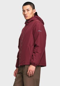 Homme portant une veste à capuche bordeaux avec fermeture éclair et poignets élastiques, et un pantalon marron, debout devant un fond clair uni.