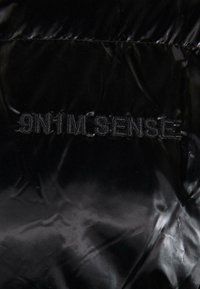 Tissu matelassé noir avec une finition brillante, comportant un texte cousu "9N1M SENSE." La texture semble lisse avec des reflets subtils.