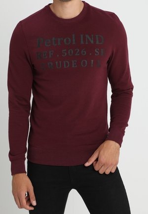 Mężczyzna w bordowej bluzie z długim rękawem z czarnym napisem „Petrol IND REF. 5026. SE CRUDE OIL” oraz czarnych spodniach.