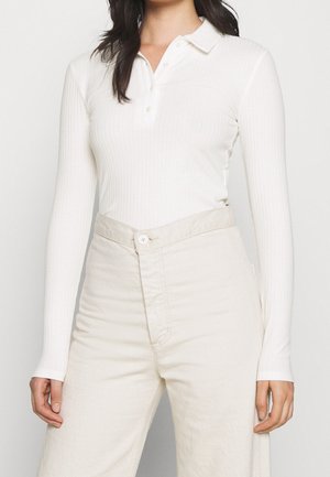 Frau trägt ein eng anliegendes weißes langärmeliges, geripptes Poloshirt, das in hoch taillierte cremefarbene Hosen gesteckt ist, vor einem einfarbigen Hintergrund.