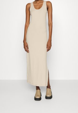 Kvinde iført en ærmeløs beige maxikjole med ribstruktur og sideslids, kombineret med chunky beige slip-on sko med sorte såler.
