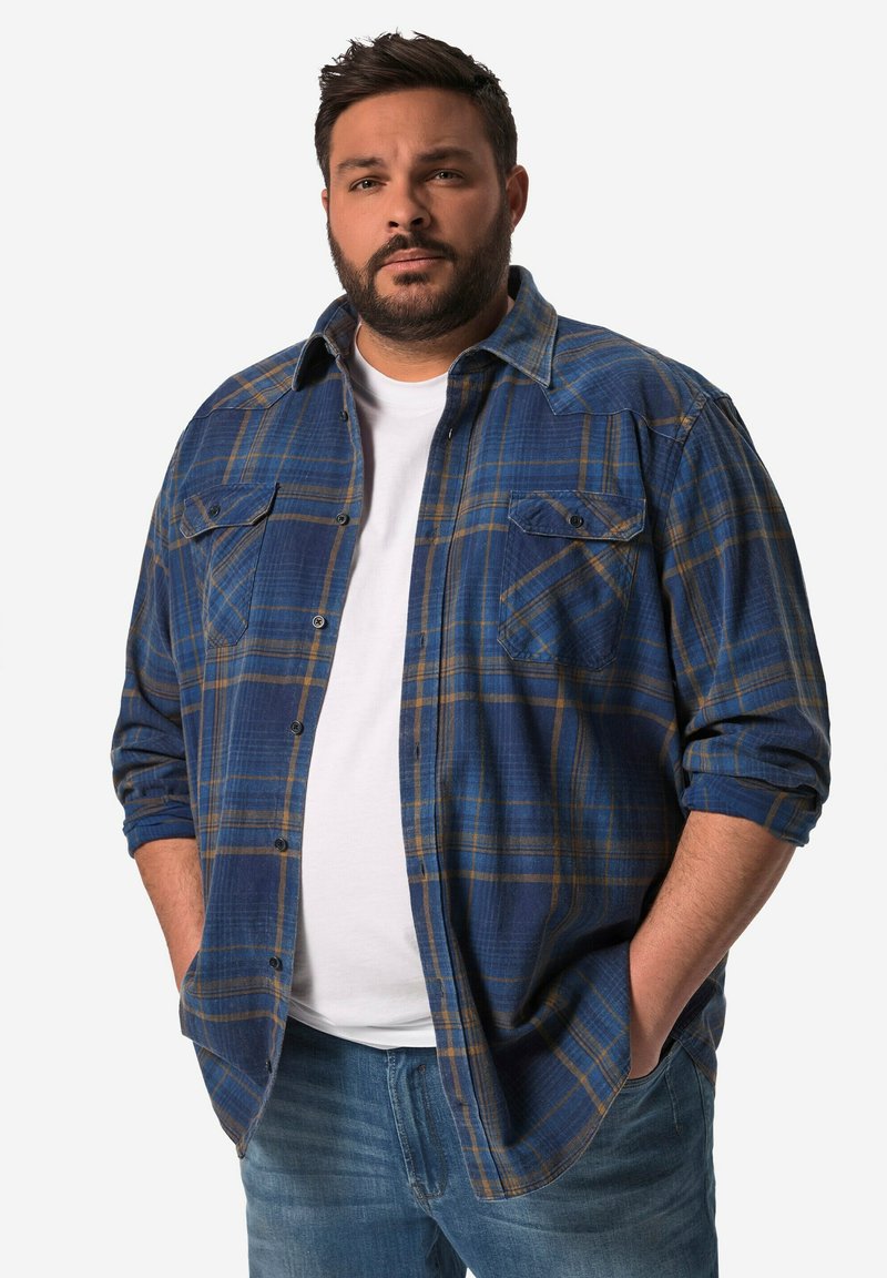 Camisa de franela a cuadros en azul con acentos amarillos, dos bolsillos en el pecho y mangas enrolladas, llevada sobre una camiseta blanca y unos vaqueros azules.