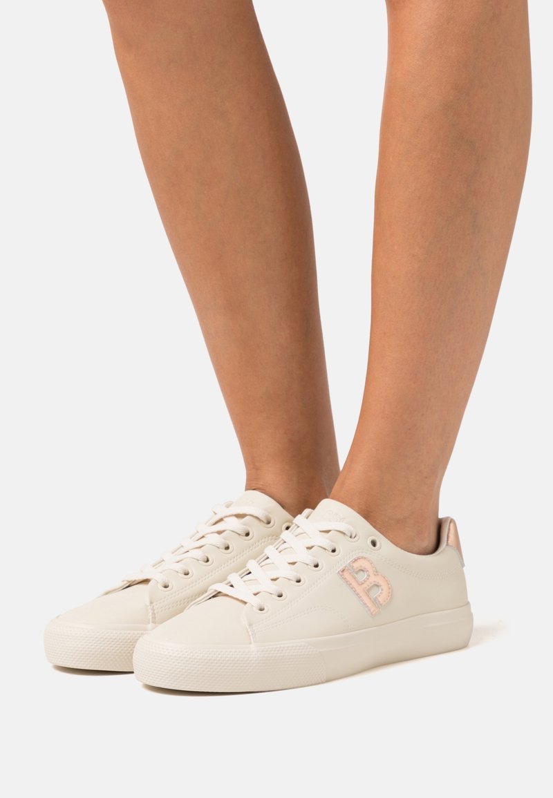 Personne portant des baskets basses blanches avec un logo "B" rose sur les côtés, debout devant un fond gris clair uni.