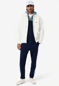 Fehér gombos dzseki kék kapucnis pulóver fölött, tengerészkék melegítőnadrággal. Világoskék részletek a nadrágon, fehér tornacipők és hozzáillő sapka.