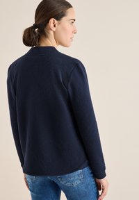 Texturovaná námořnicky modrá bunda s jemným geometrickým vzorem, s hladkým povrchem a kulatým výstřihem, kombinovaná se světle modrými džínami.