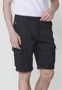Shorts cargo noirs avec un devant plat, un ourlet roulé et de multiples poches latérales. Fabriqués en tissu durable avec une texture lisse.