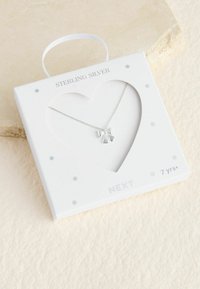 Halskæde i sterlingsølv med en sløjfevedhæng, vist i en hvid hjerteformet æske med grå prikker og et båndhåndtag.