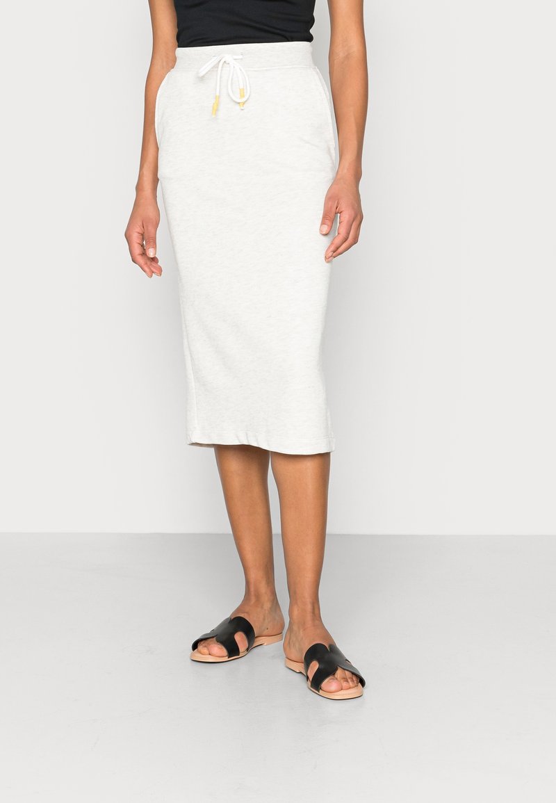 Jupe en coton gris clair, mi-longue, avec une taille élastique et un cordon de serrage. Associée à des sandales plates noires. Texture lisse, design décontracté.