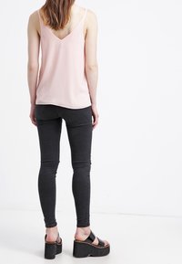 Femme se tenant de dos, portant un top sans manches rose pâle, un jean skinny noir et des sandales compensées noires.