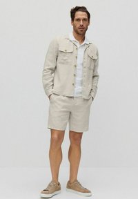 Vaaleanbeige pellavapusero, jossa napit ja kaksi rintataskua, yhdistettynä samanvärisiin shortseihin. Alla valkoinen polo-paita ja ruskeat lenkkarit.