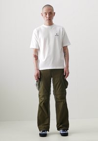 T-shirt blanc à manches courtes avec un petit logo, associé à un pantalon cargo vert olive avec des poches latérales et une coupe décontractée. Chaussures noires.