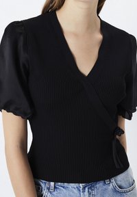 Femme portant un haut cache-cœur noir côtelé avec des manches courtes bouffantes en satin et un jean en denim bleu clair sur un fond blanc