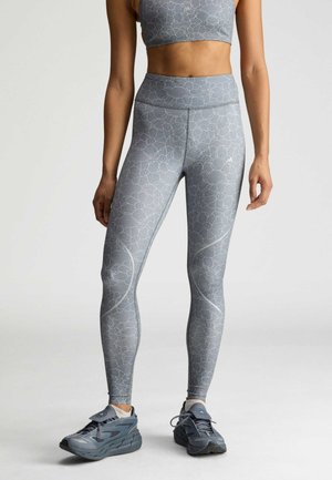 Vrouw die grijze leggings met een bloemenpatroon en hoge taille draagt, bijpassend crop top en grijze sportschoenen, staand tegen een effen achtergrond.