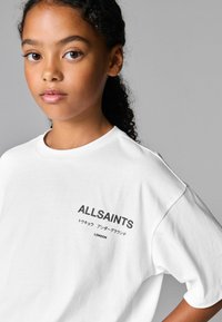 Bílý krátký rukávový tričko s tištěným logem "ALLSAINTS" a černým textem na přední straně, s kulatým výstřihem a uvolněným střihem.