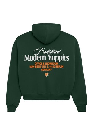 Jeune femme portant un sweat à capuche vert foncé, un short gris à fines rayures jusqu’au genou, des chaussettes blanches, des mocassins noirs et une casquette de baseball blanche, posant sur fond blanc.