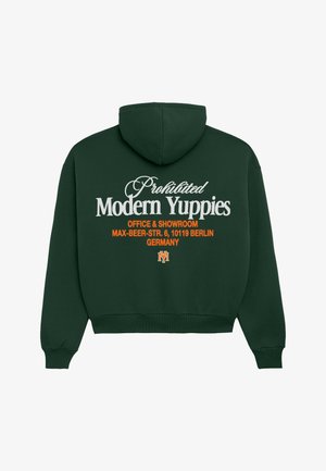 Młoda kobieta ubrana w ciemnozieloną bluzę z kapturem, szare spodnie w paski za kolano, białe skarpetki, czarne mokasyny i białą czapkę z daszkiem, pozująca na białym tle.