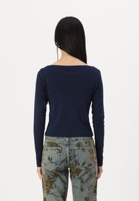 Jūros mėlynos spalvos ilgomis rankovėmis marškiniai su kvadratiniu iškirpimu, derinami su šviesiai mėlynomis džinsais, kurie turi rudų gėlių raštų. Lygus medvilnės audinys.